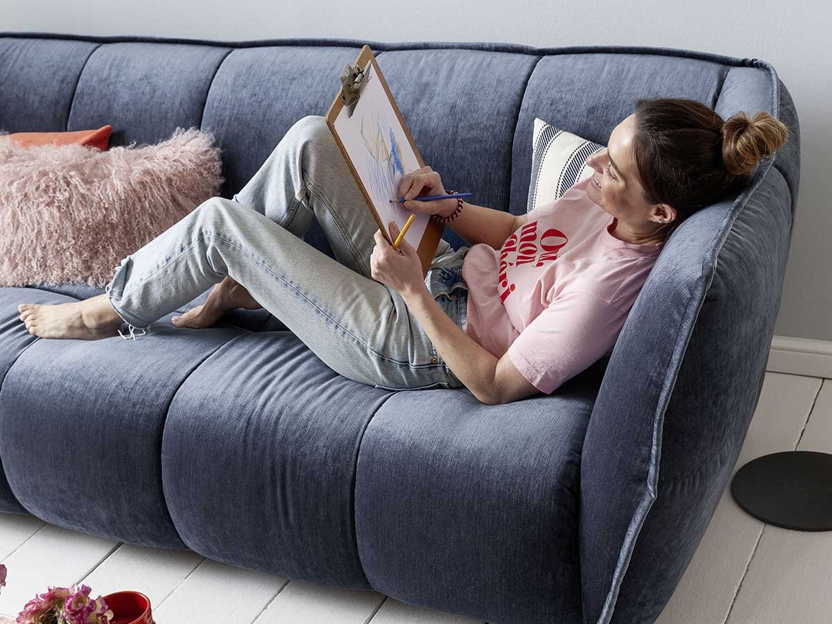 Frau liegt auf blauem Sofa und zeichnet mit Buntstiften ein Bild auf weißem Papier